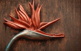 A red, blue-tinted and bronze metal Bird of Paradise flower on a brown wood-grained metal surface.