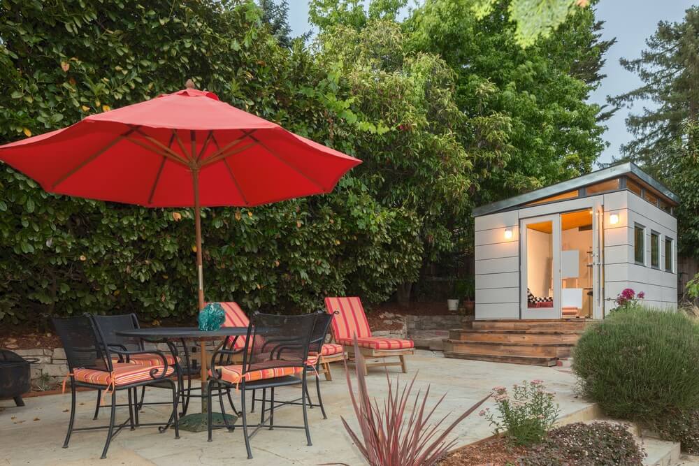 A backyard studio with outdoor dining table and red shade umbrella in the foreground